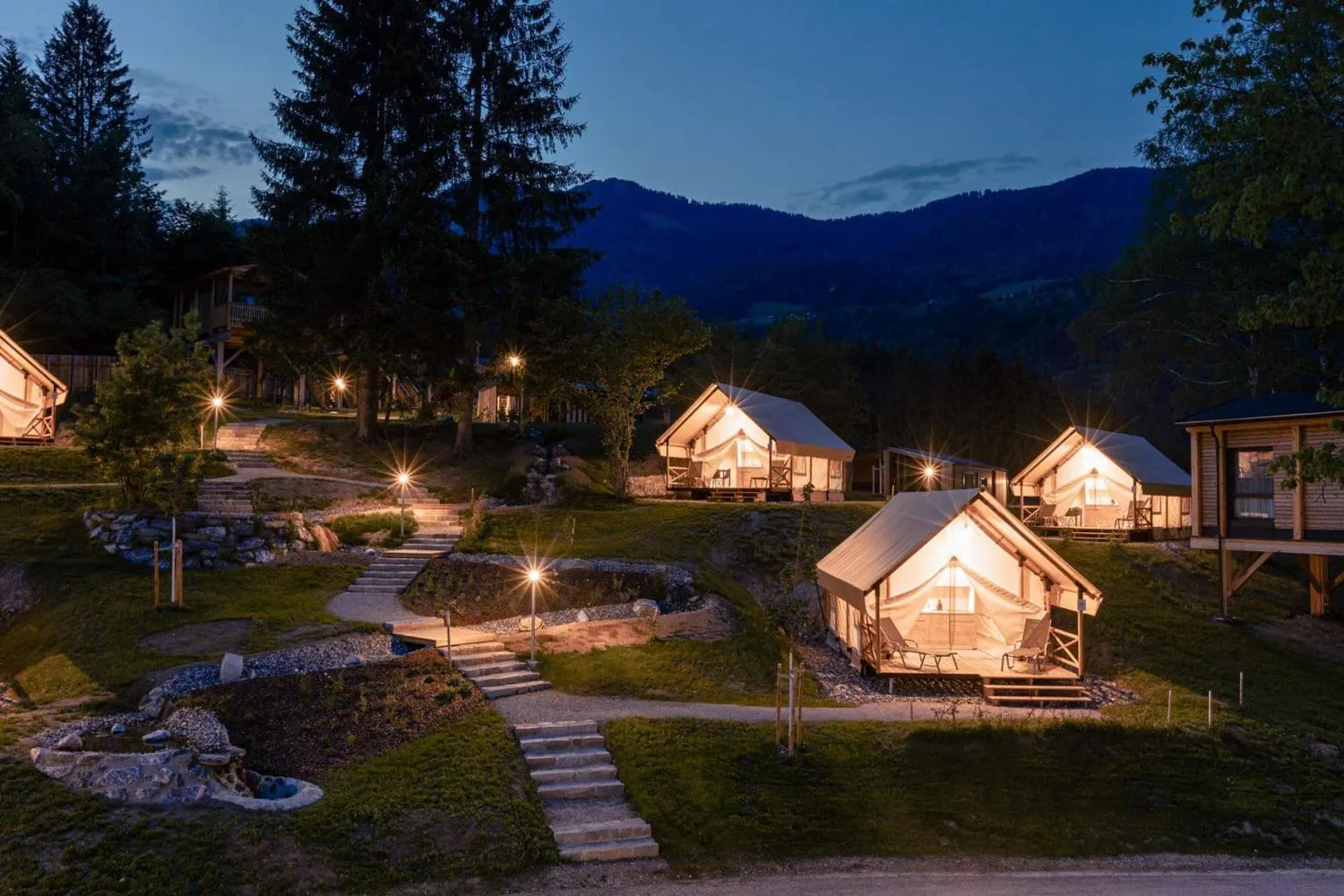 Beleuchtete Glamping-Pods nachts mit Feuergrube und Bergen