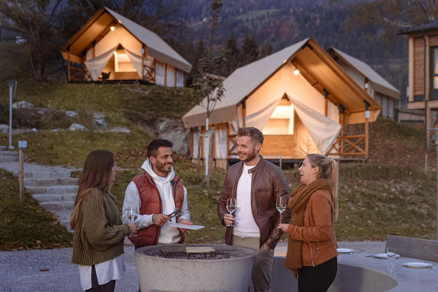 Vier Personen am Feuer mit beleuchteten Glamping-Pods im Hintergrund