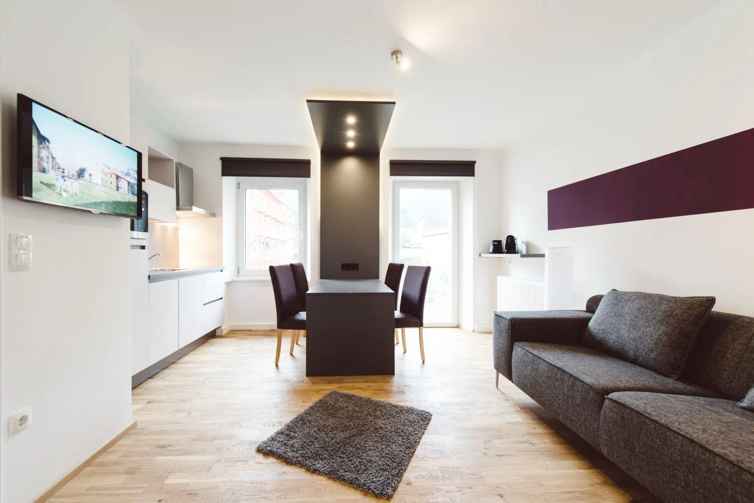 Wohnbereich mit grauem Sofa, Essplatz mit braunen Stühlen und TV an Wand
