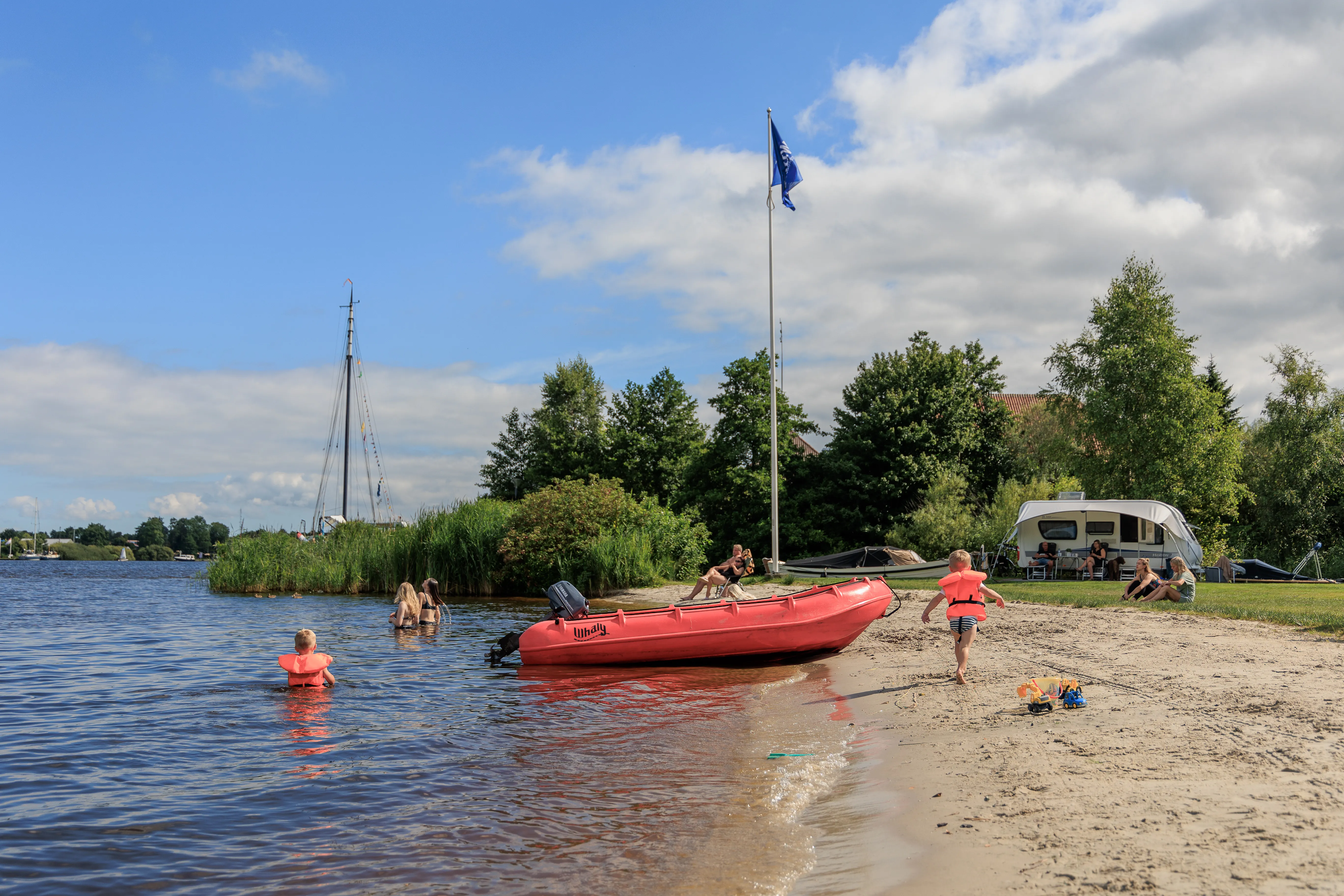 Sandstrand am Wasser mit rotem Schlauchboot, Kindern in Schwimmwesten und einer Fahne am Mast