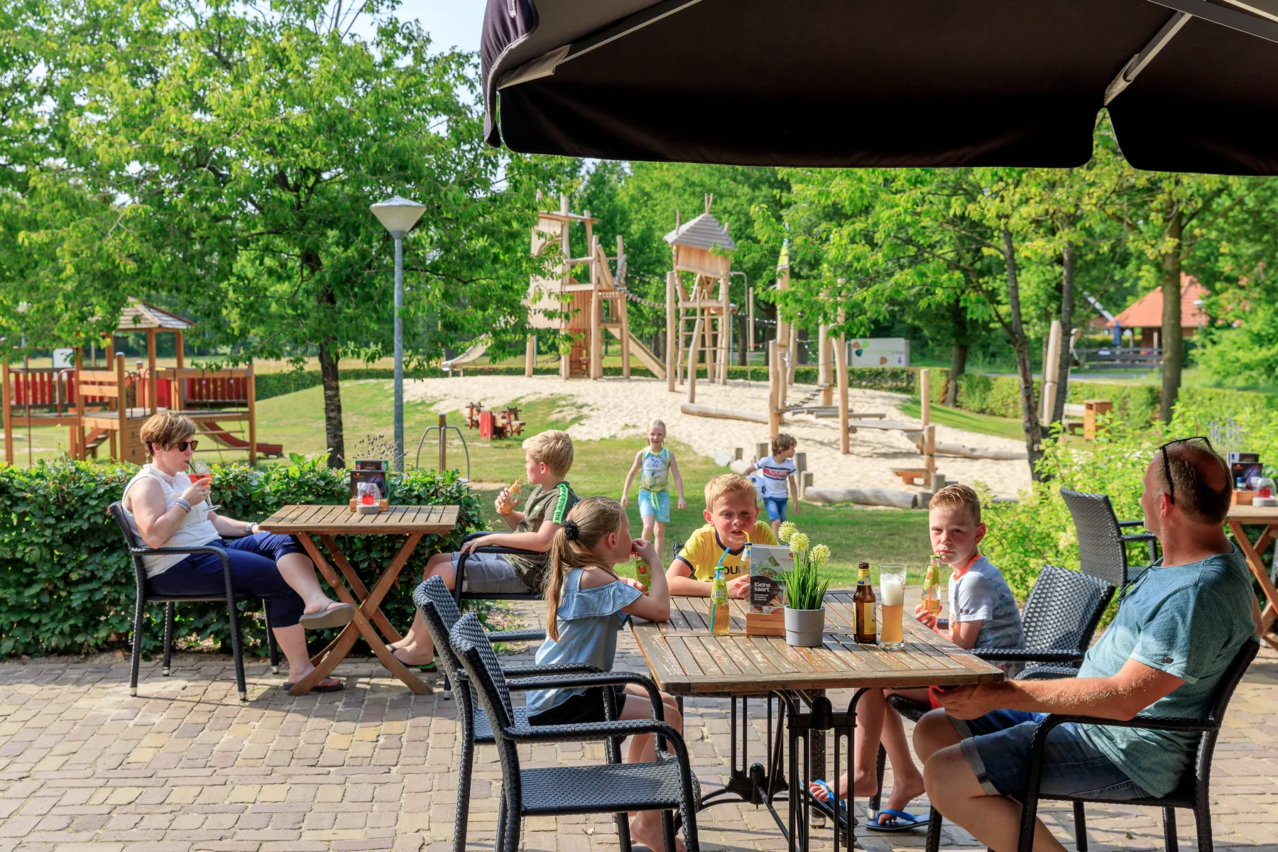 Gäste an Tischen auf der Restaurantterrasse unter Sonnenschirmen, dahinter der Holzspielplatz