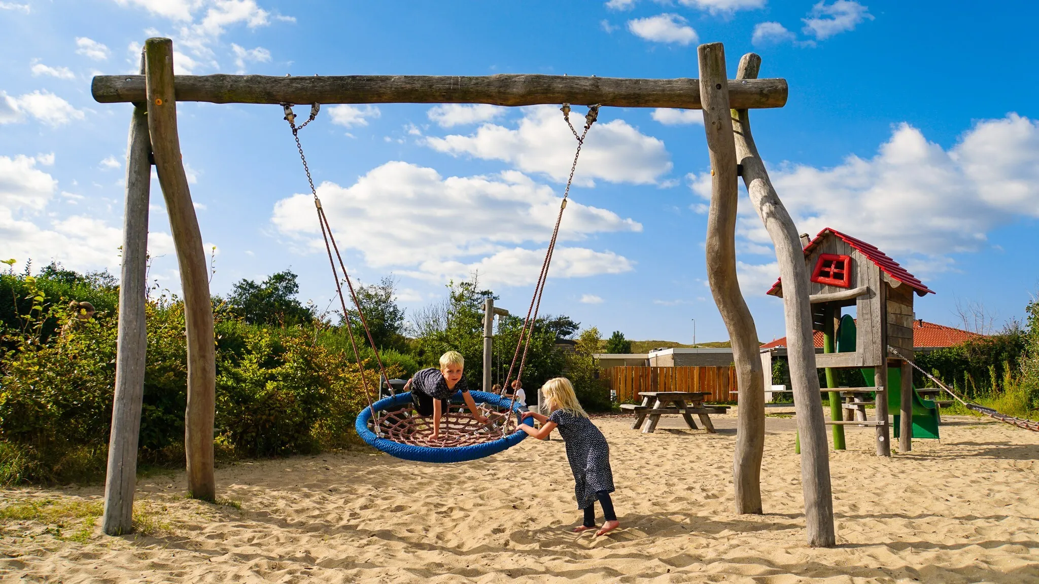 Zwei Kinder an der Nestschaukel auf dem Sandspielplatz, dahinter ein Spielhaus mit Rutsche