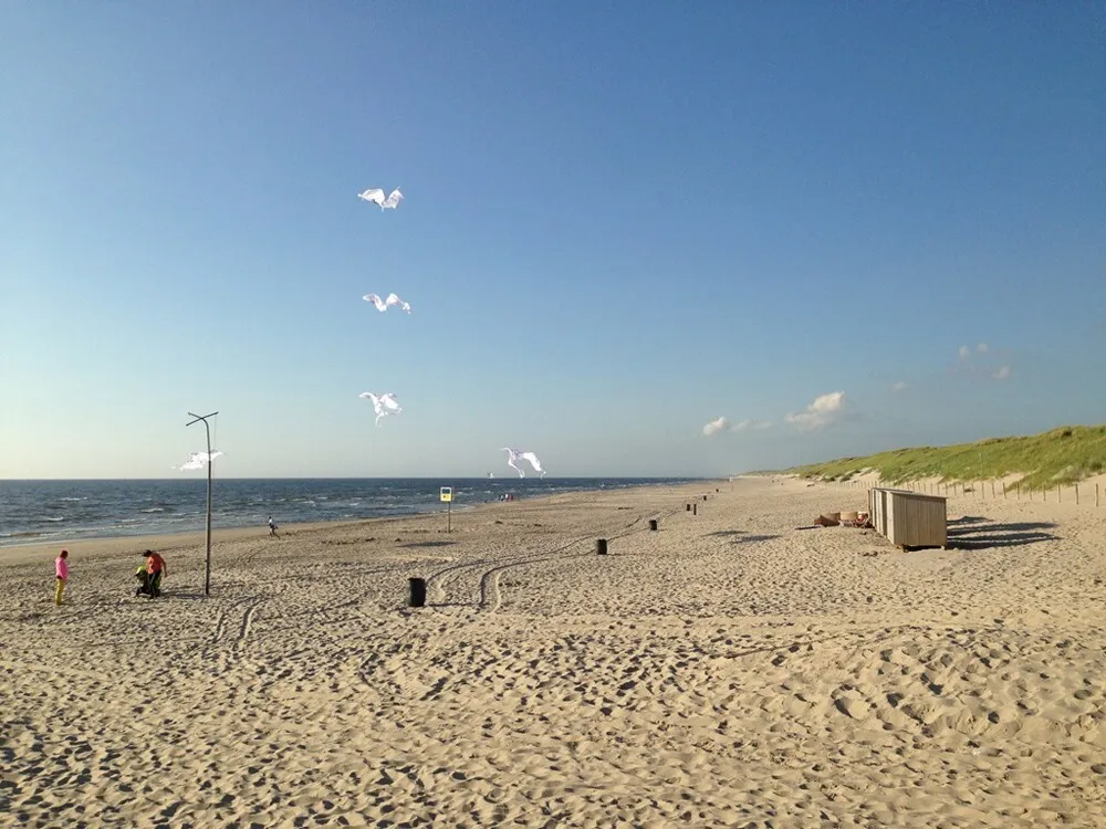 Breiter Sandstrand mit Lenkdrachen über dem Wasser und Dünen am rechten Rand