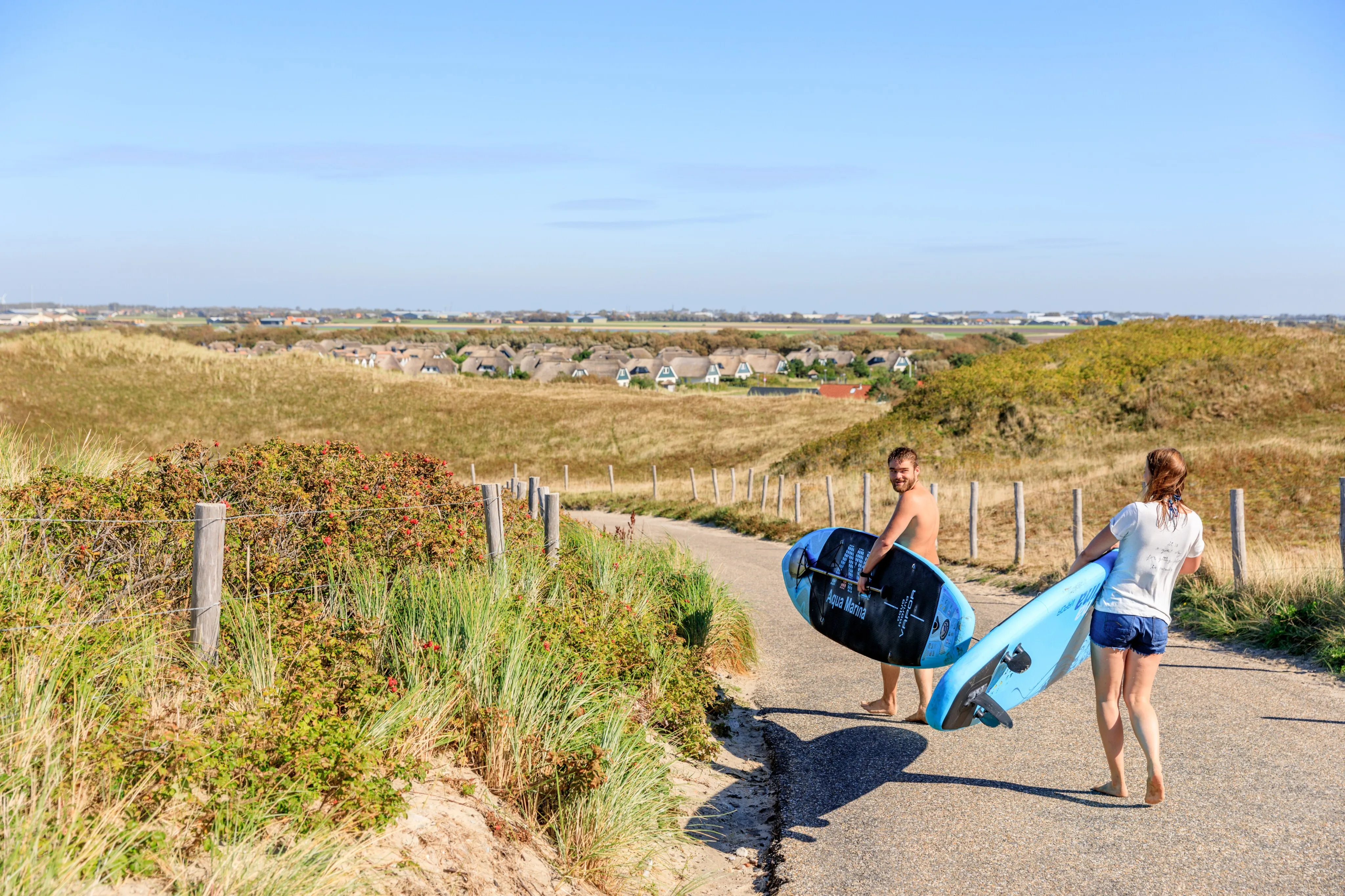 Zwei Personen tragen Surfbretter über den Dünenweg, im Hintergrund reetgedeckte Häuser