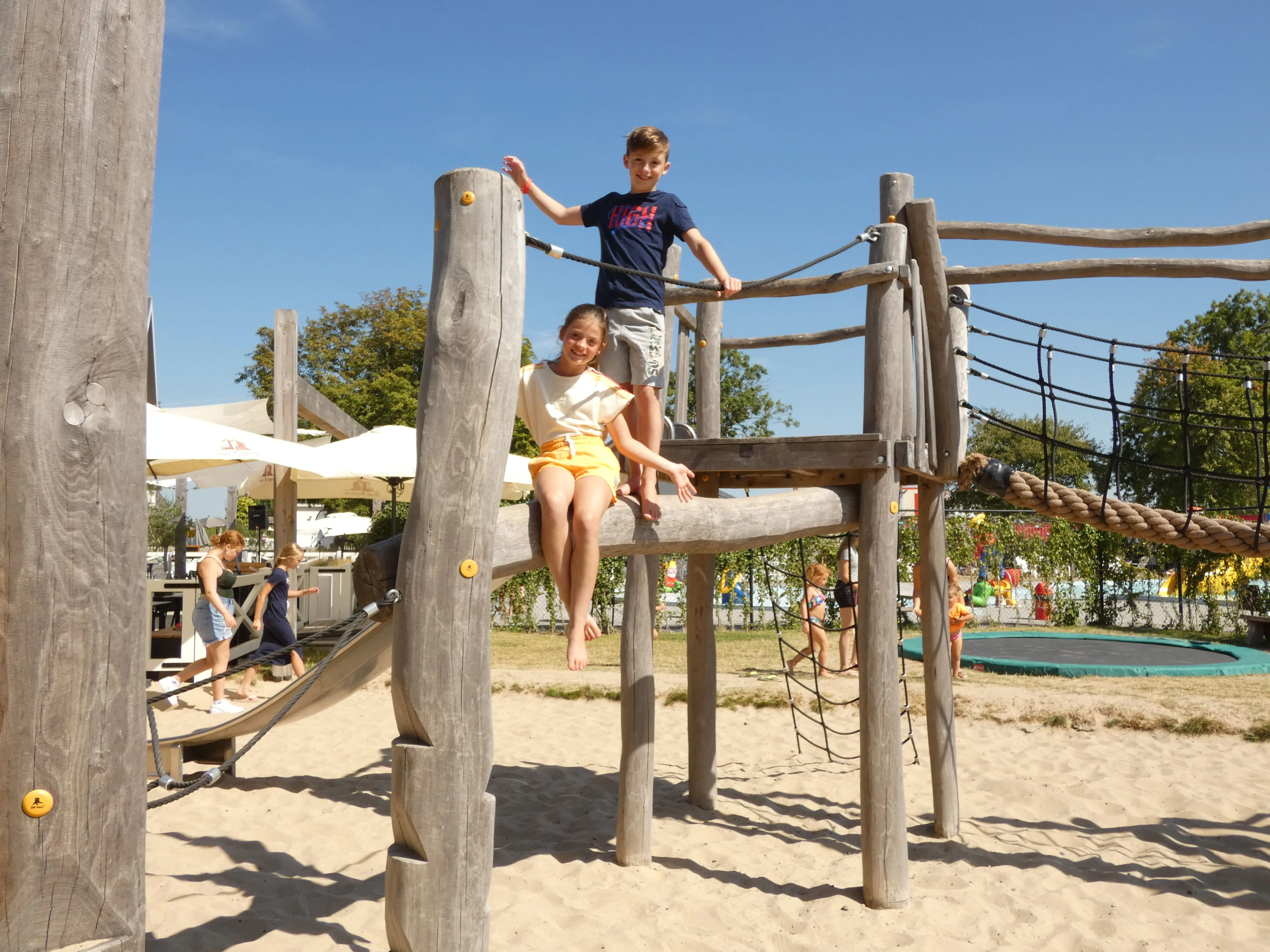 Kinder auf einem Holzklettergerüst im Sand, dahinter Trampolin und Sonnenschirme
