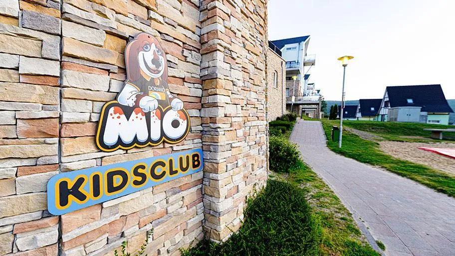 Kidsclub-Eingangsschild mit Mio-Maskottchen an Natursteinmauer
