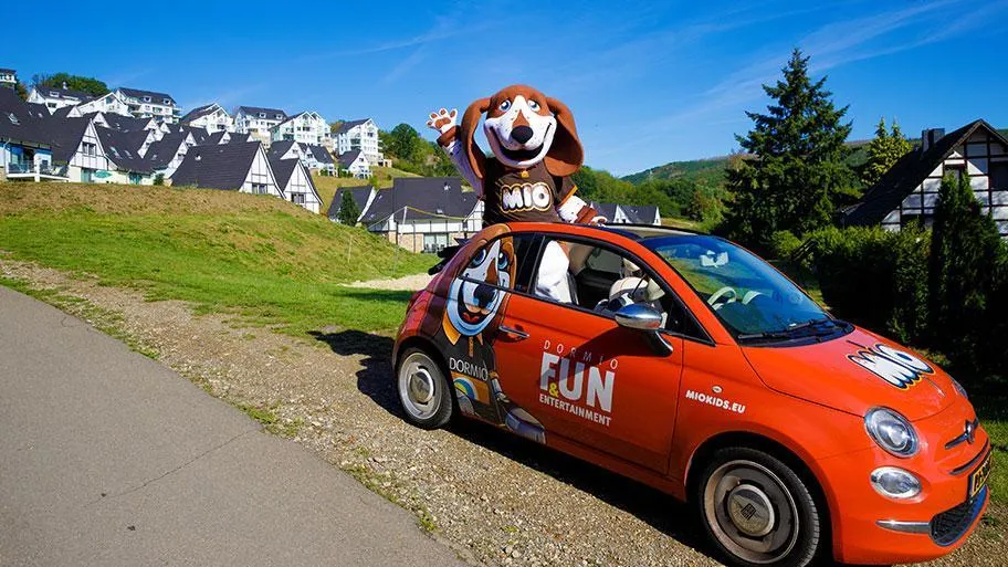 Rotes Kinderspielzeugauto mit Mio-Maskottchen vor Ferienpark-Häusern