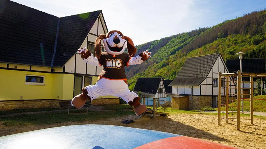 Mio-Hundmaskottchen springt vor Ferienpark-Häusern und Eifelhügeln