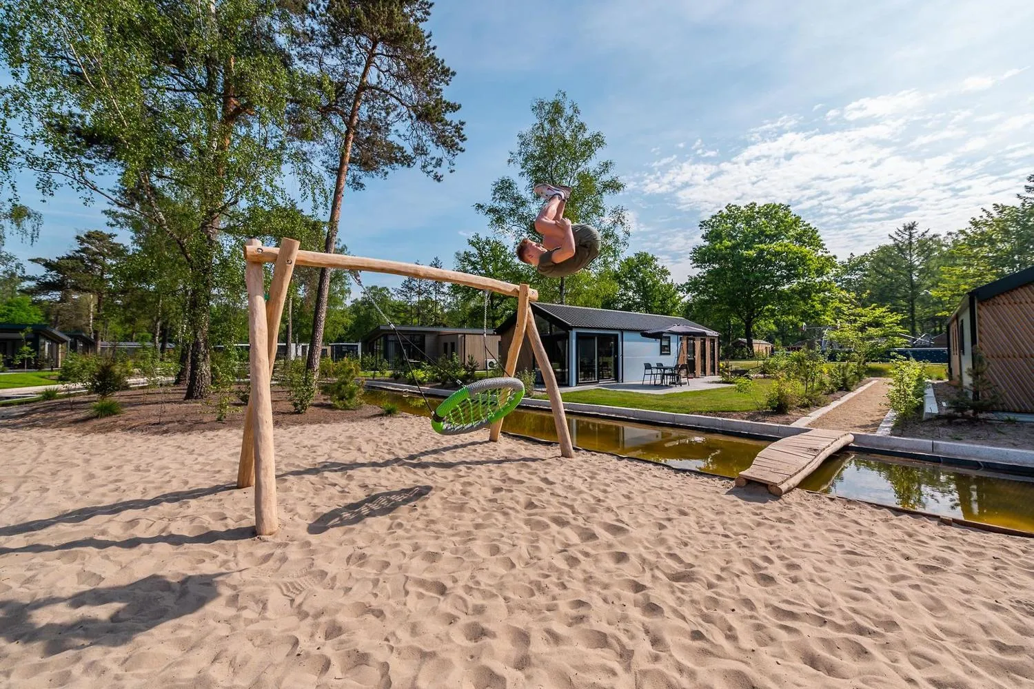 Spielplatz mit Holzschaukel auf Sand, moderne Ferienhäuser und Kanal dahinter