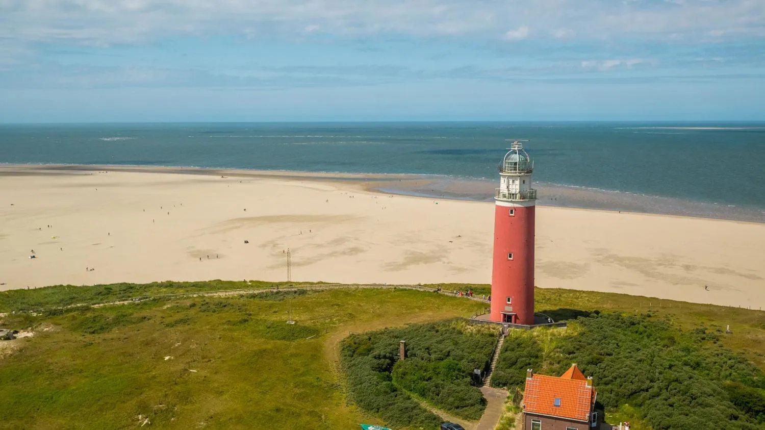 Roter Leuchtturm auf grüner Düne über breitem Sandstrand