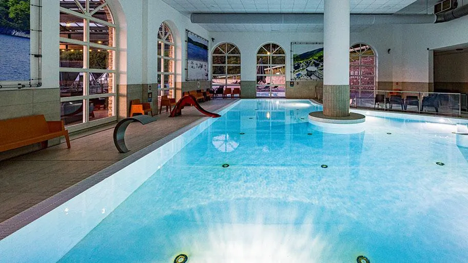 Modernes Hallenschwimmbad mit hellem Wasser, großen Fenstern und Lounge-Bereich